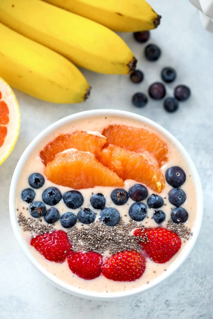 Overhead view of grapefruit smoothie bowl topped with grapefruit sections, blueberries, chia seeds, and sliced strawberries with bananas, blueberries, and grapefruit half in the background