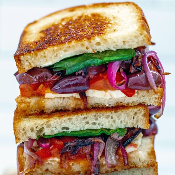 Closeup of two halves of a Greek grilled cheese sandwich stacked on each other with feta, red peppers, red onion, kalamata olives, tomato, and spinach peeking out