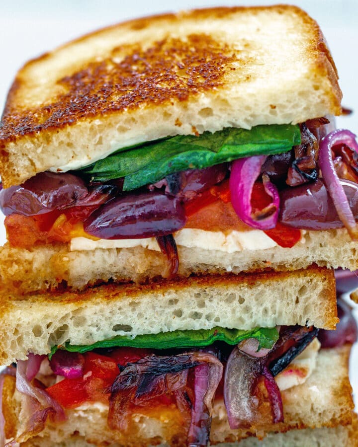 Closeup of two halves of a Greek grilled cheese sandwich stacked on each other with feta, red peppers, red onion, kalamata olives, tomato, and spinach peeking out