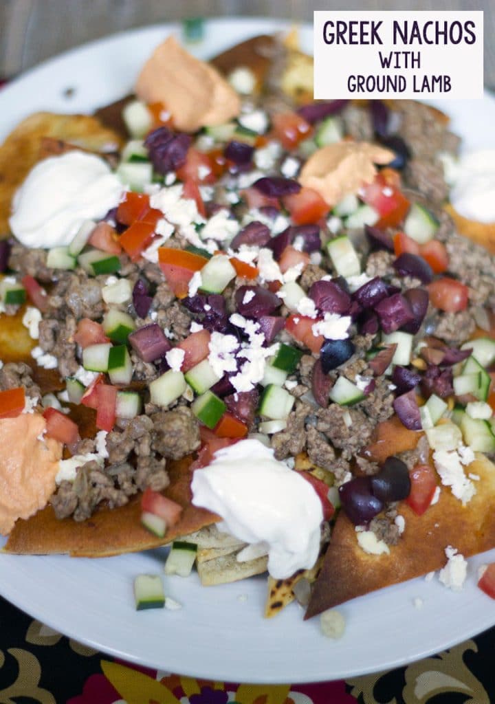 Overhead view of a plate of Greek nachos with ground lamb, feta cheese, cucumbers, tomatoes, olives, hummus, and yogurt with recipe title at the top of the image