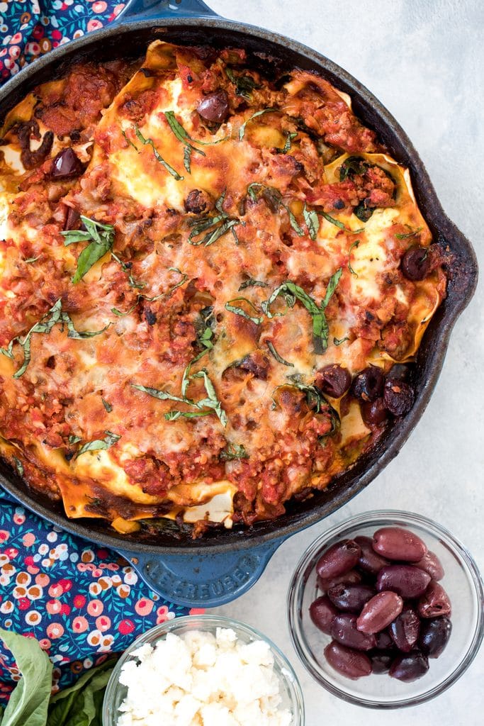 Overhead view of Greek lasagna with ground lamb and kalamata olives in a skillet with small bowls of olives and feta in background