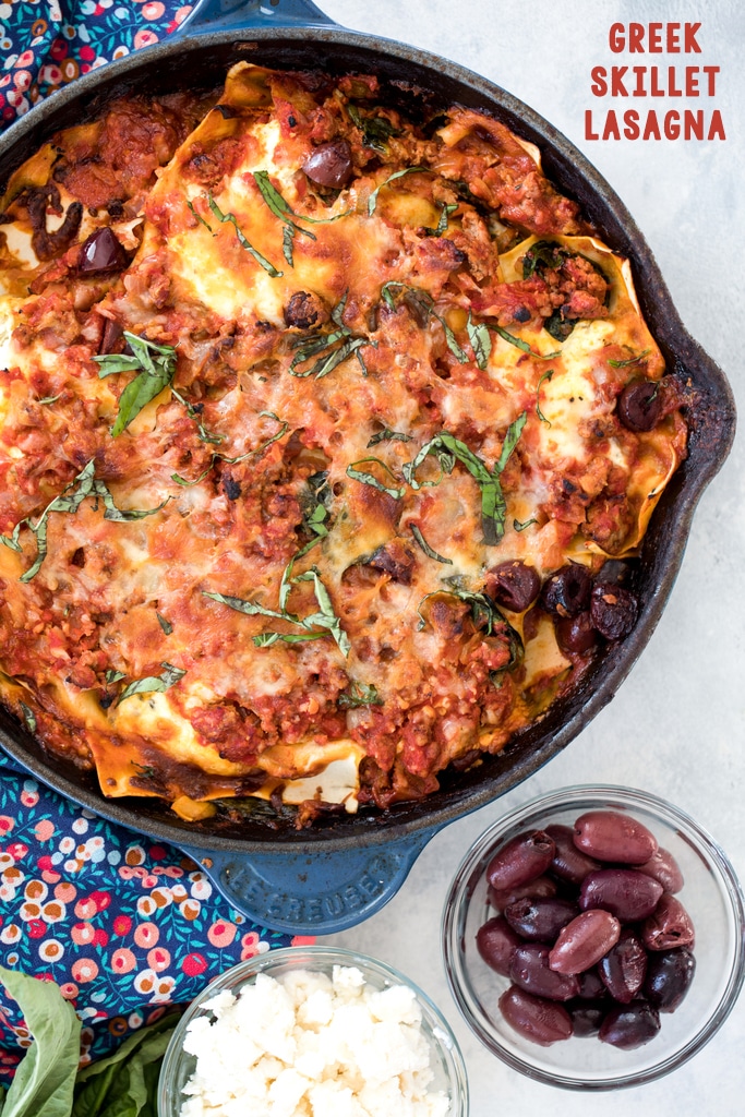 Overhead view of Greek lasagna with ground lamb and kalamata olives in a skillet with small bowls of olives and feta in background and recipe title at top