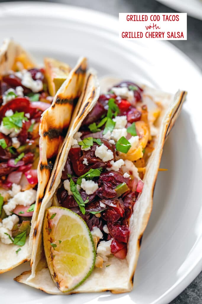 Overhead closeup view of grilled cod tacos with cherry salsa, cheese, scallions, onions, and lime with recipe title at top