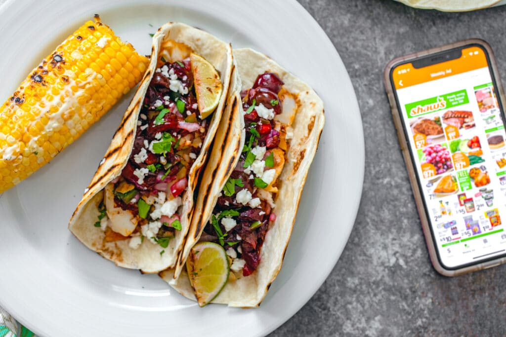 Bird's eye view of two grilled cod tacos with grilled cherry salsa with side of corn on the cob with iPhone open to Shaw's Supermarket app on the side