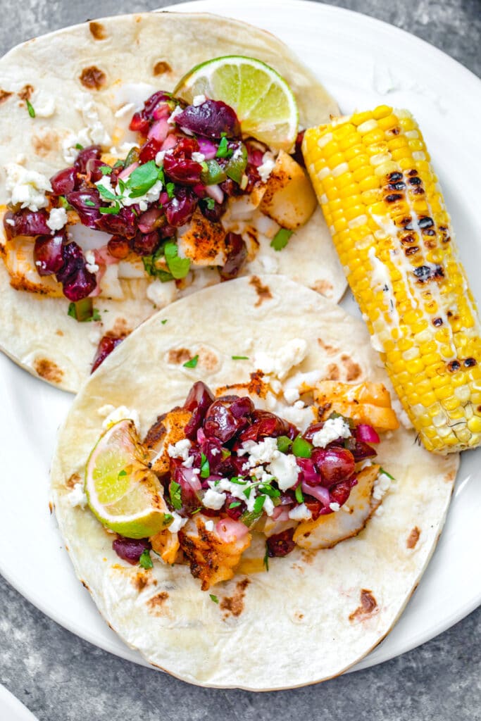 Overhead view of two open-faced grilled cod tacos topped with cherry salsa with side of corn on the cob