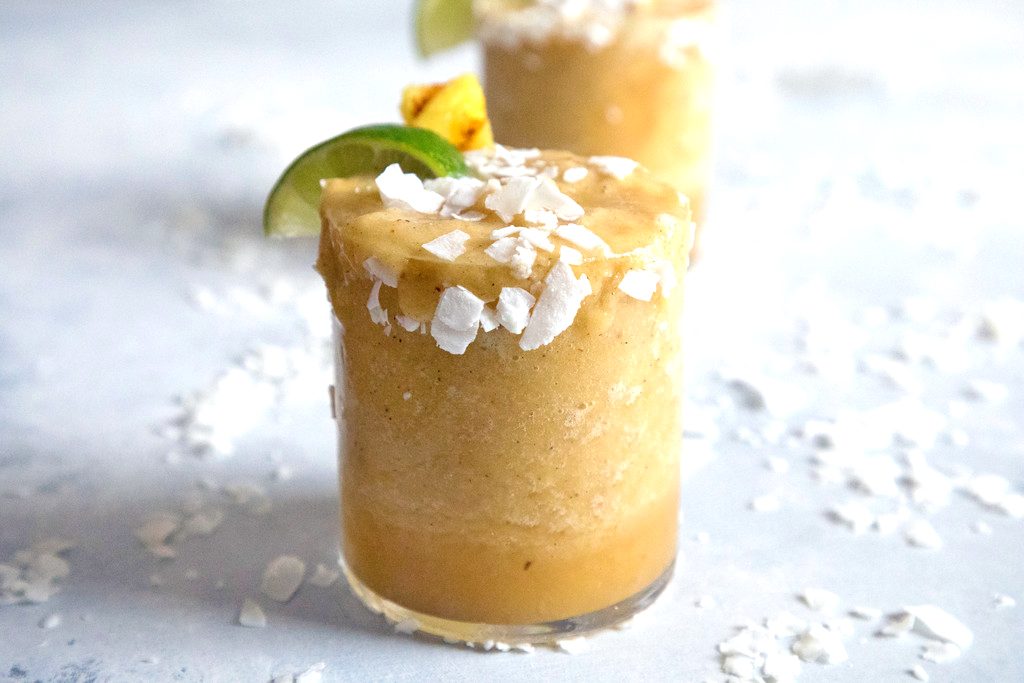 Landscape closeup view of a grilled pineapple coconut margarita with coconut, lime wedge, and pineapple garnish with coconut sprinkled around and second cocktail in the background