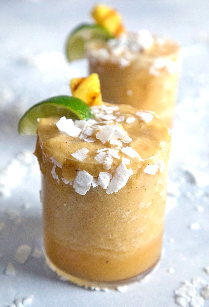 Head-on closeup view of grilled pineapple coconut margarita with coconut, lime wedge, and pineapple garnish with coconut sprinkled around and second cocktail in background