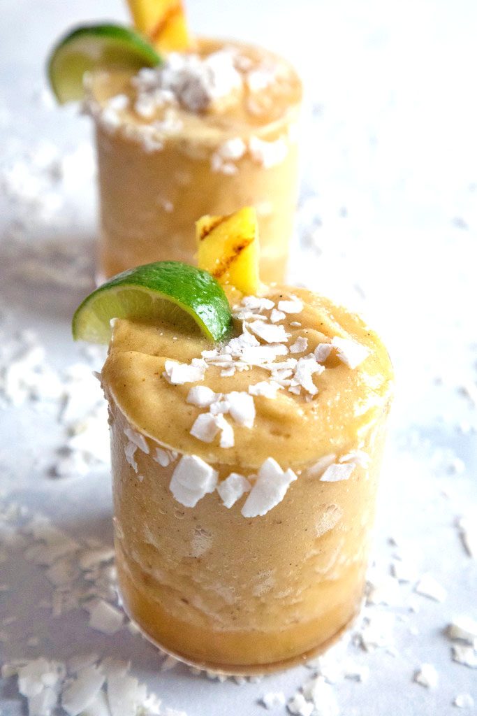 Head-on view of grilled pineapple coconut margarita with coconut, lime, and pineapple garnish with second cocktail in background