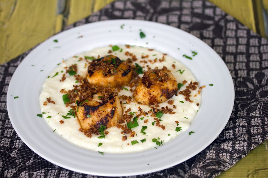 Landscape view of a white bowl of cheesy grits topped with grilled scallops and panko