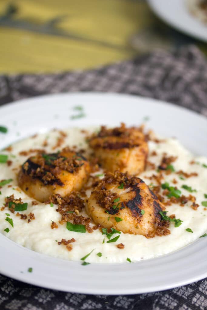 Head-on view of a white bowl of cheesy grits topped with grilled scallops and panko