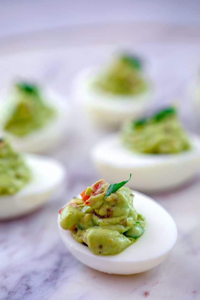 Head-on close-up of guacamole deviled egg on marbled surface with more eggs in the background