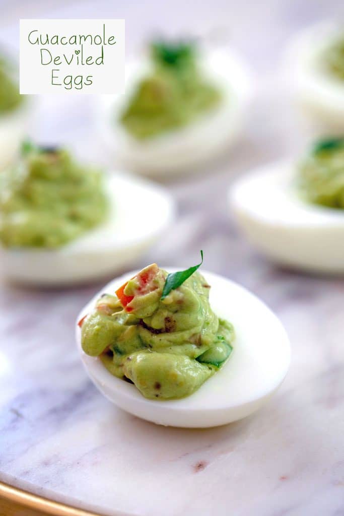 Head-on view of guacamole deviled egg on marble surface with other eggs in the background and "Guacamole Deviled Eggs" text at top