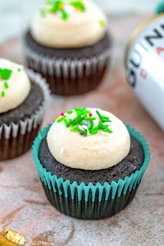 Head-on view of a Guinness cupcake with Guinness frosting and green sprinkles with more cupcakes and can of Guinness in the background.