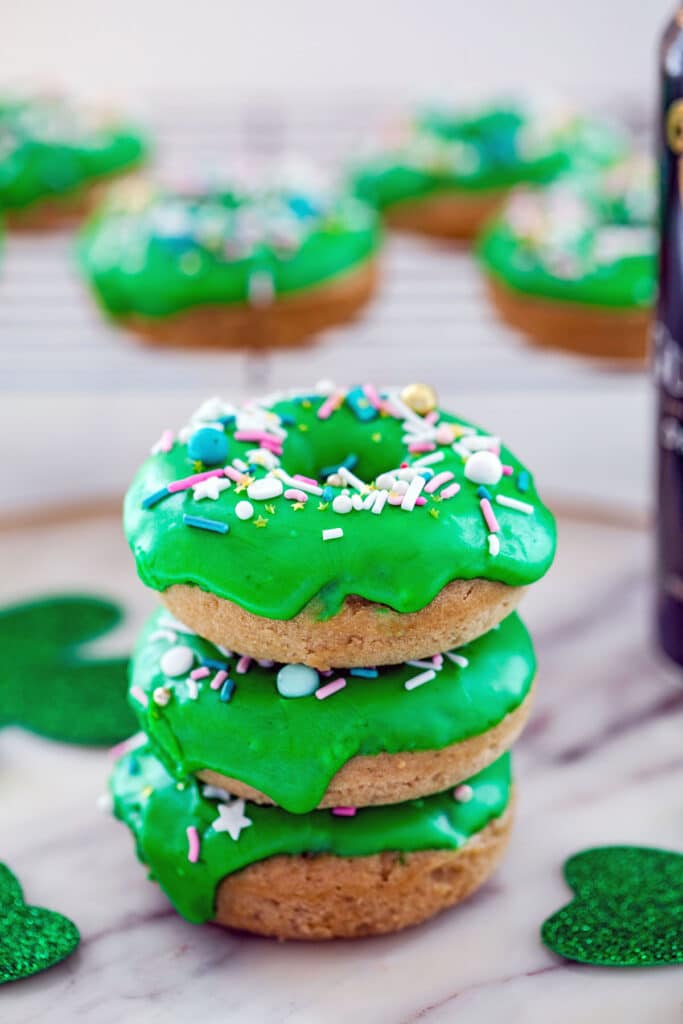 Head-on closeup view of three Guinness doughnuts stacked with green frosting and sprinkles and baking rack with more doughnuts in the background with glitter shamrocks.