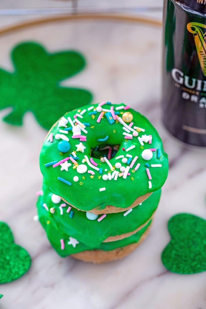 Overhead view of a stack of Guinness doughnuts with bright green Irish whiskey frosting and sprinkles on a marble surface with glitter shamrocks and can of Guinness beer in the background