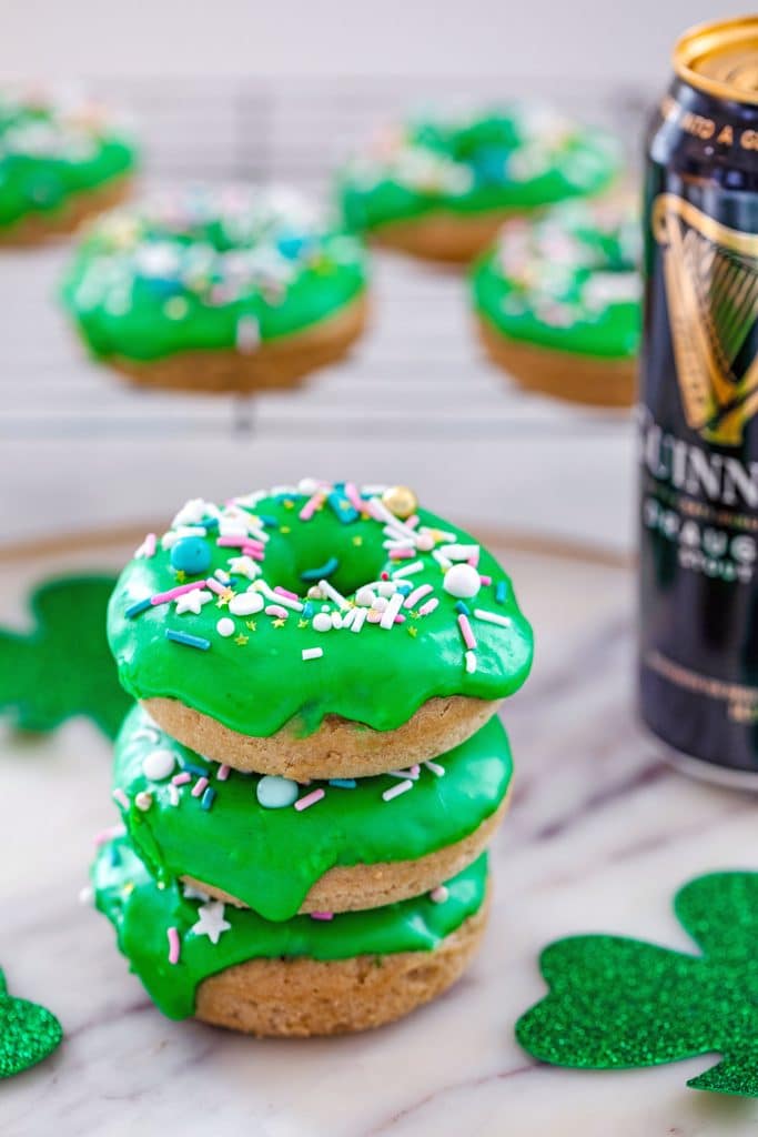 Stack of four Guinness doughnuts with baking rack with more doughnuts in the background, a tall can of Guinness beer and glitter shamrocks on marble surface