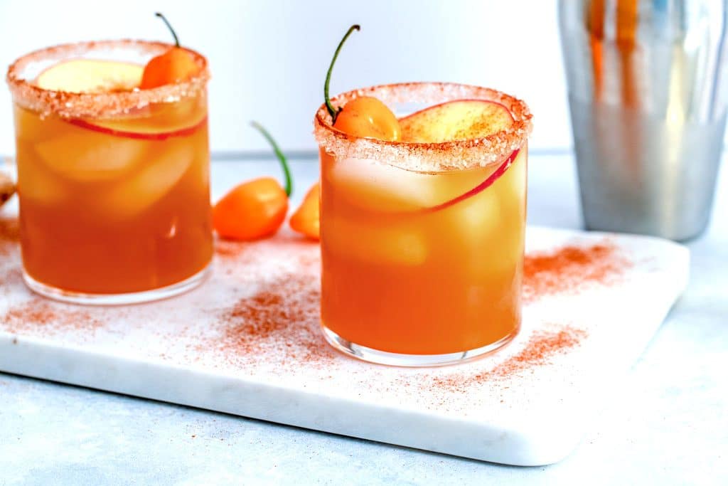 Landscape view of two habanero ginger apple cider cocktails on a marble surface sprinkled with cinnamon with habanero peppers and cocktail shaker in the background