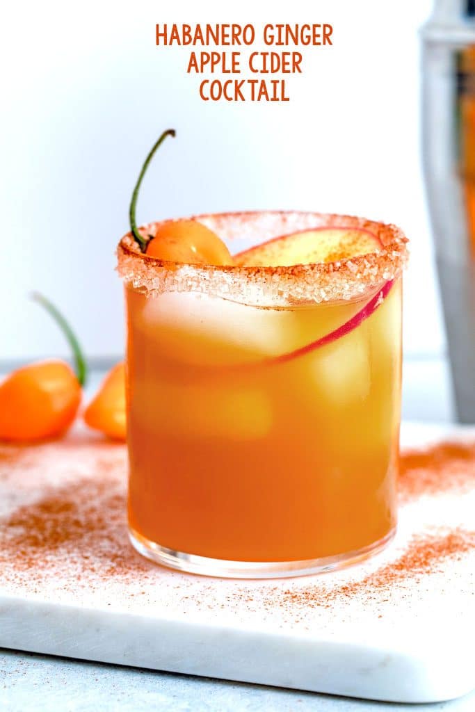 Head-on image of habanero ginger apple cider cocktail with apple and habanero garnish on marble board sprinkled with cinnamon and recipe title at the top of the image
