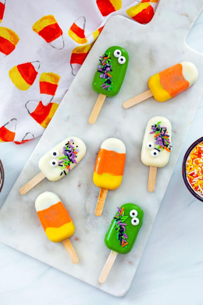 Bird's eye view of Halloween cakesicles (candy corn, ghosts, and green monsters) on a marble slab with candy corn tea towel in background