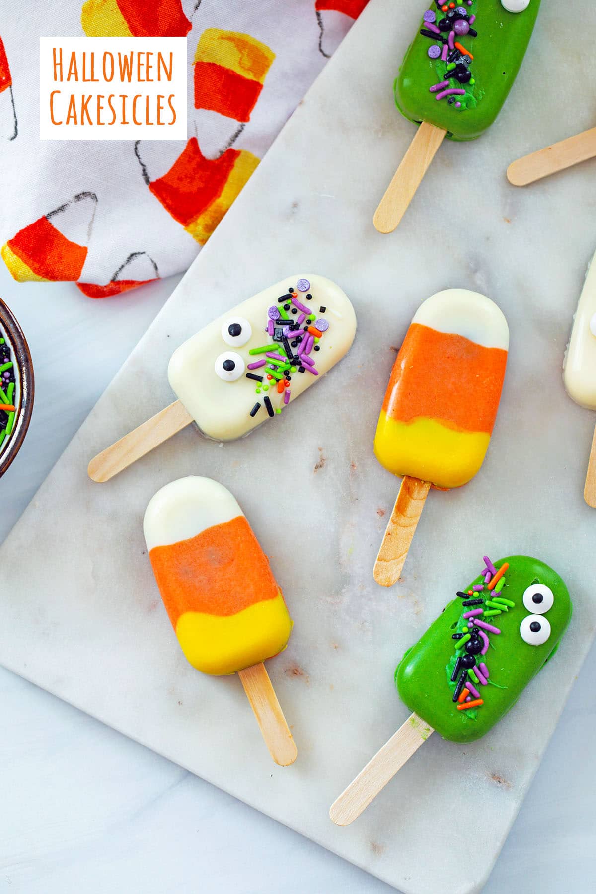 Overhead view of various Halloween Cakesicles on marble board, including candy corn cakesicles and ghost cakesicle with recipe title at top.