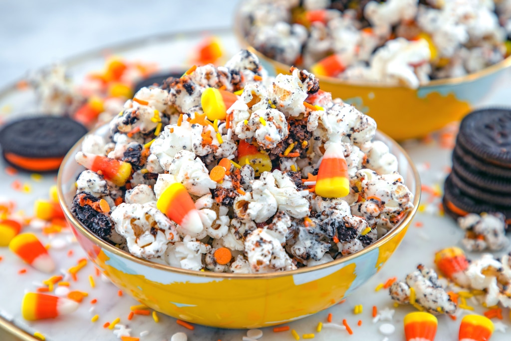 Landscape head-on close-up view of a bowl of Halloween party popcorn with candy corn, Halloween sprinkles, and Halloween Oreo cookies all around with second bowl of popcorn in the background.