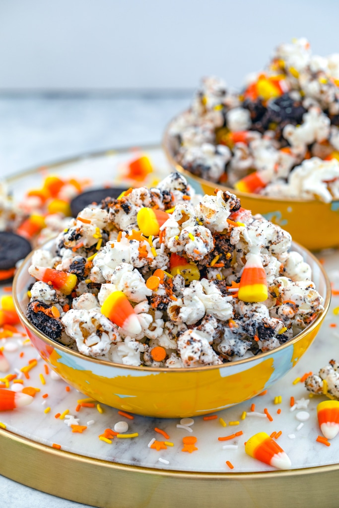 Head-on view of a bowl of Halloween party popcorn on a marble tray with Halloween sprinkles and Oreo cookies and candy corn all around with second bowl of popcorn in the background.