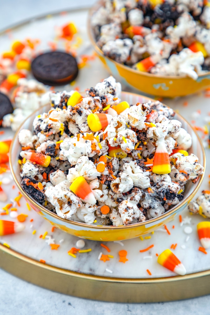 Overhead view of a bowl of Halloween party popcorn with candy corn, orange and yellow sprinkles, and Halloween Oreo cookies all around and a second bowl of popcorn in the background.