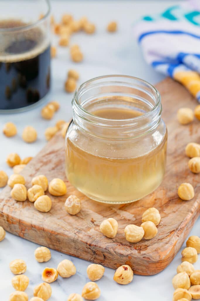Small jar of hazelnut syrup with hazelnuts all around and cup of coffee in the background.
