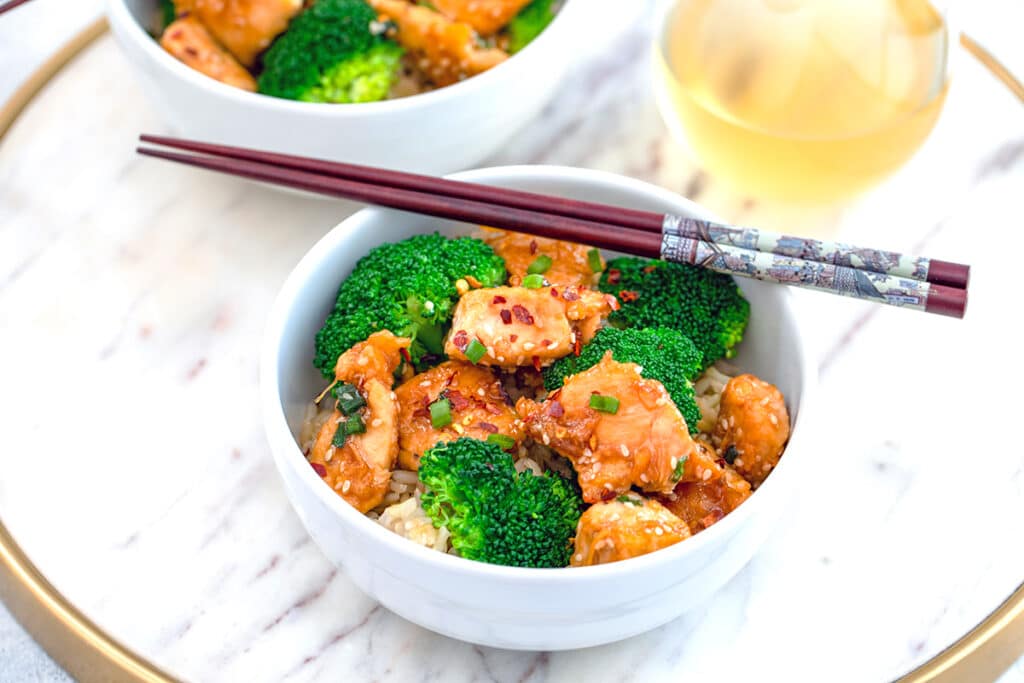 Landscape view of a bowl of healthier sesame chicken with broccoli and brown rice with chopsticks.