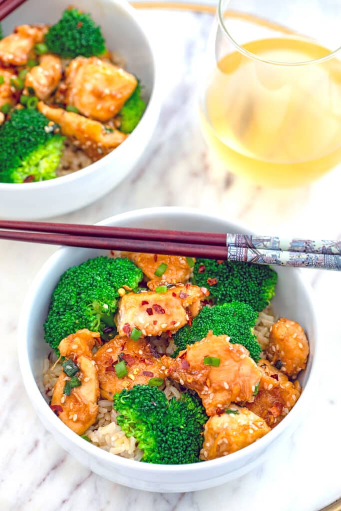 Lighter sesame chicken in a bowl with rice and broccoli with second bowl in background.