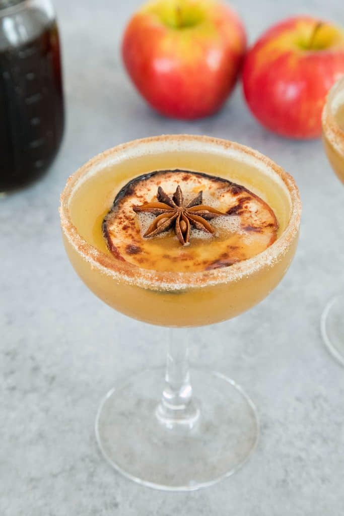 View of a holiday spiced honeycrisp apple cocktail in a coup glass with a dried apple and star anise garnish with jar of simple syrup and apples in the background