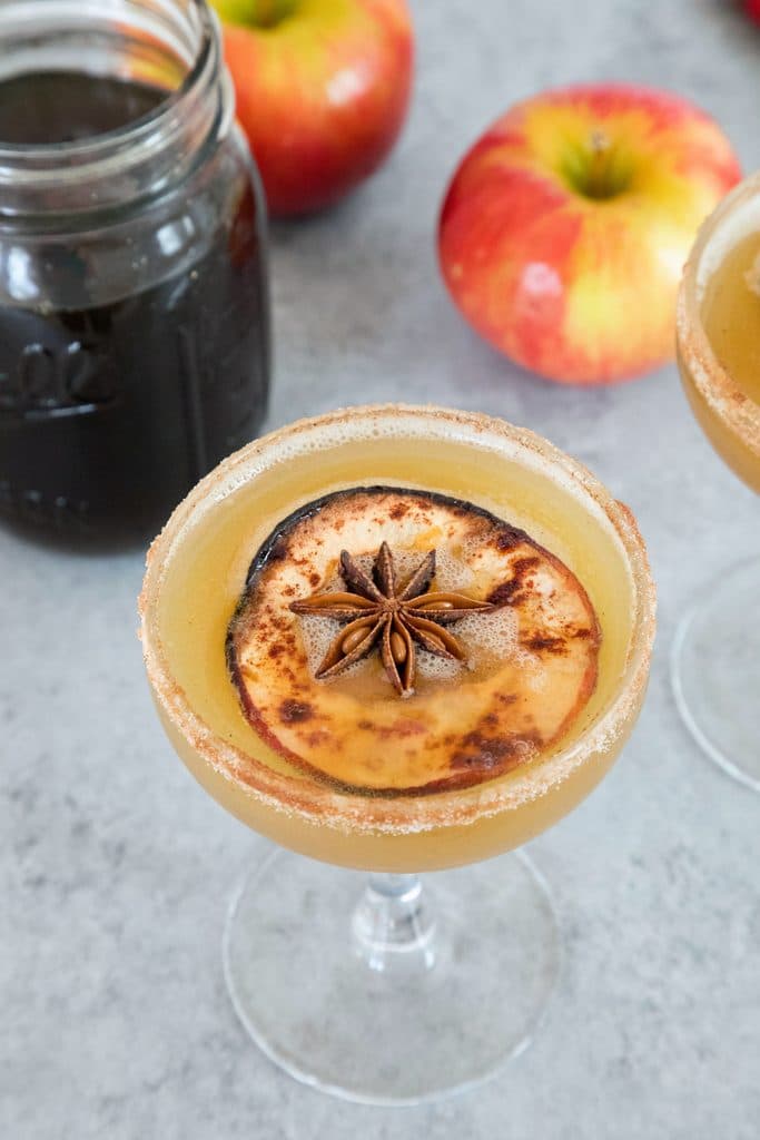 Overhead closeup view of a holiday spiced honeycrisp apple cocktail in a coup glass with jar of simple syrup and apples in the background