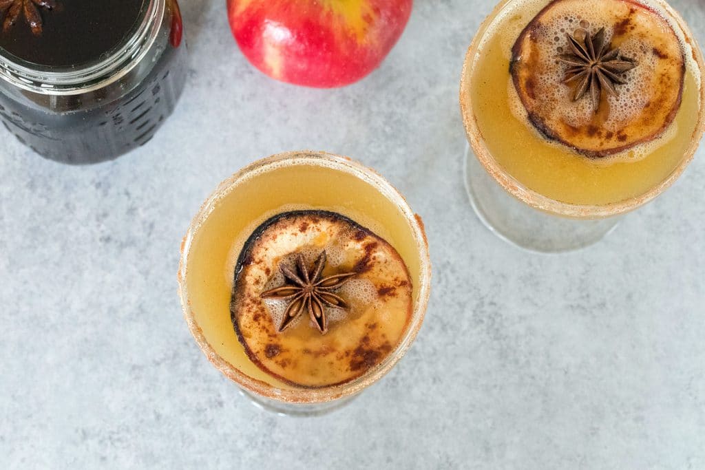 Overhead landscape view of two holiday spiced honeycrisp apple cocktails with jar of simple syrup and apple