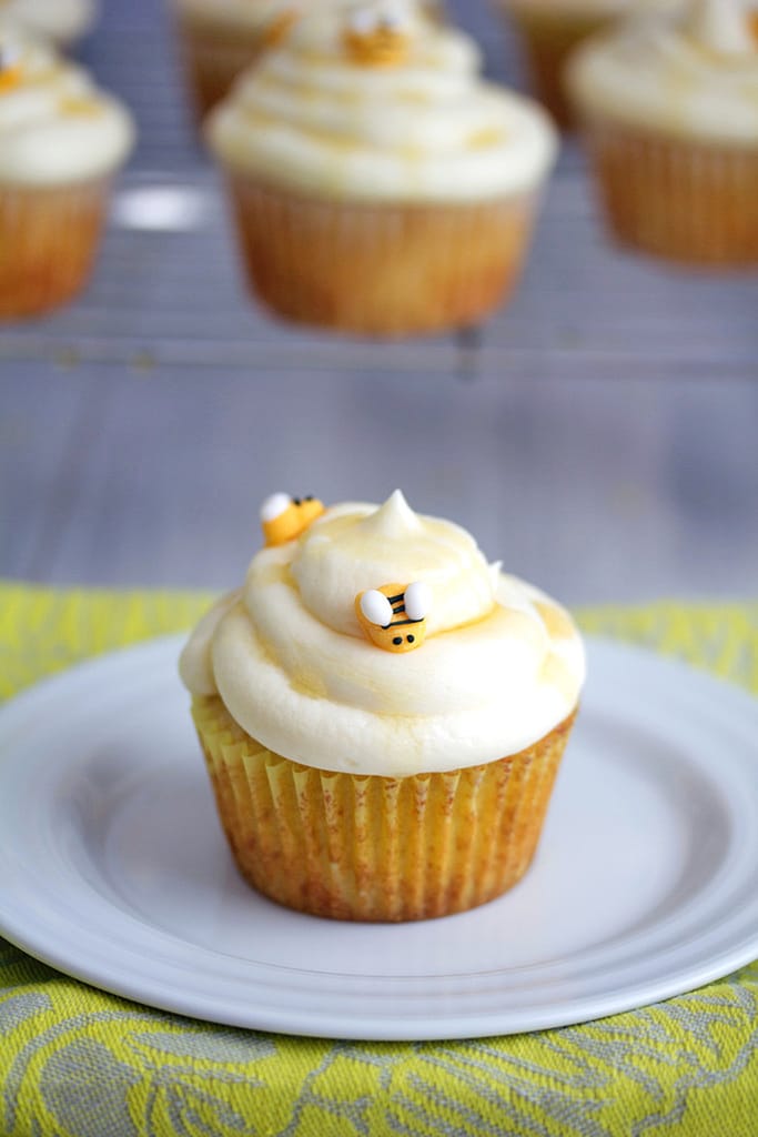 Head on view of a honey cupcake topped with honey buttercream icing with honey drizzle and little honeybee decorations with more cupcakes on rack in background and recipe title at top.