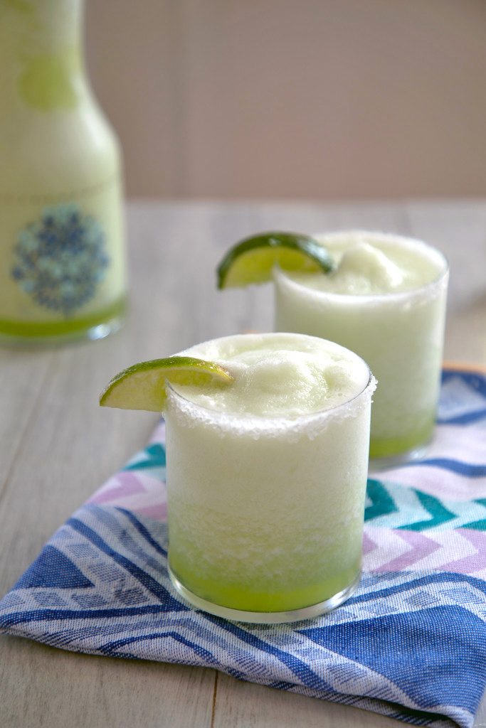 Head-on view of two bright green honeydew margaritas with lime wedge garnish on a colorful napkin with pitcher of margaritas in the background