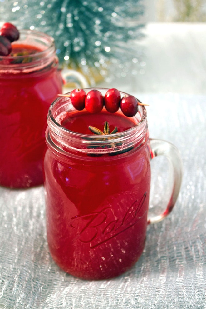 A mason jar filled with hot spiced cranberry cocktail with star anise and cranberry garnish with a second cocktail and tinsel in the background