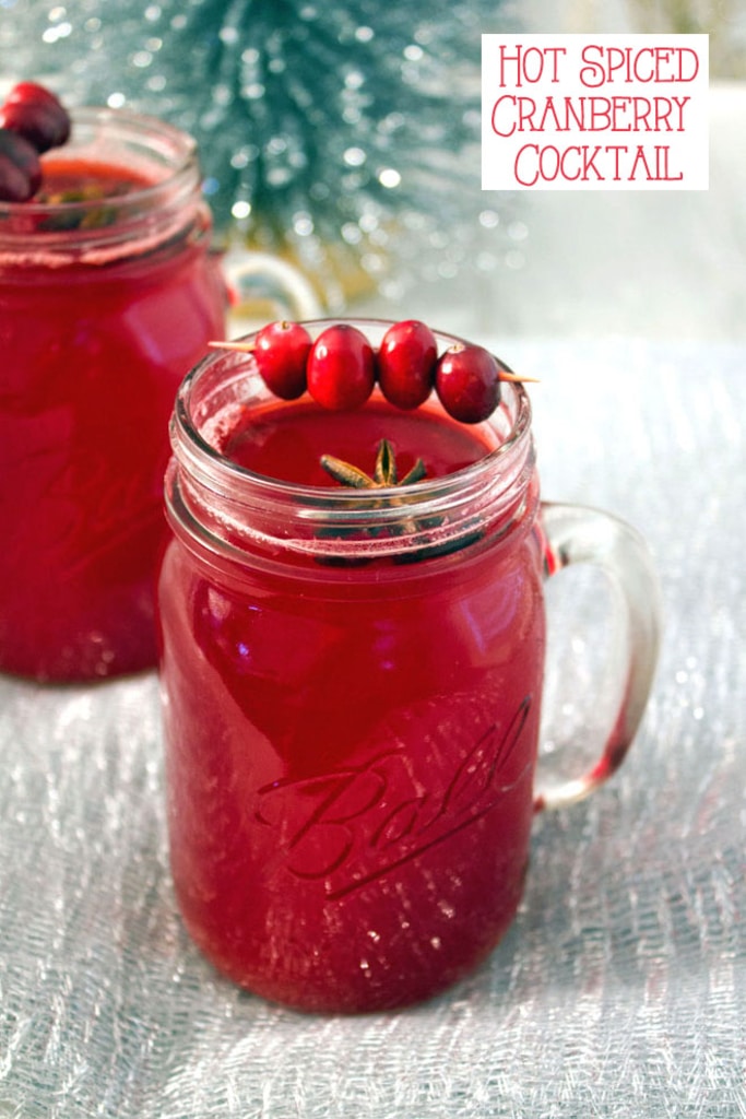 A mason jar filled with hot spiced cranberry cocktail with star anise and cranberry garnish with a second cocktail and tinsel in the background with recipe title at top