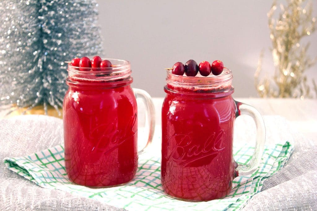 A landscape photo featuring two mason jars of hot spiced cranberry cocktails with cranberry garnishes and a small tinsel tree in the background