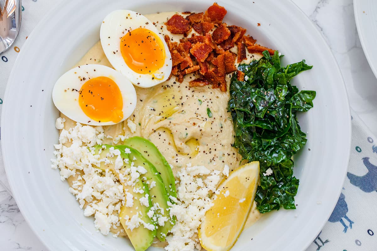 Landscape overhead closeup view of hummus breakfast bowl with sliced avocado, kale, feta, halved soft-boiled egg, crumbled bacon, and lemon wedge.