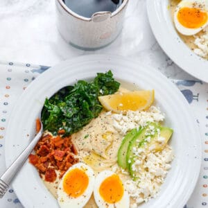 Overhead view of hummus breakfast bowl with halved soft-boiled egg, crumbled bacon, feta cheese, sliced avocado, kale, and lemon wedge with cup of coffee and second hummus bowl in the background