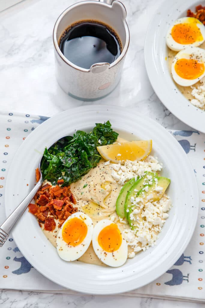 Overhead view of hummus breakfast bowl with halved soft-boiled egg, crumbled bacon, feta cheese, sliced avocado, kale, and lemon wedge with cup of coffee and second hummus bowl in the background