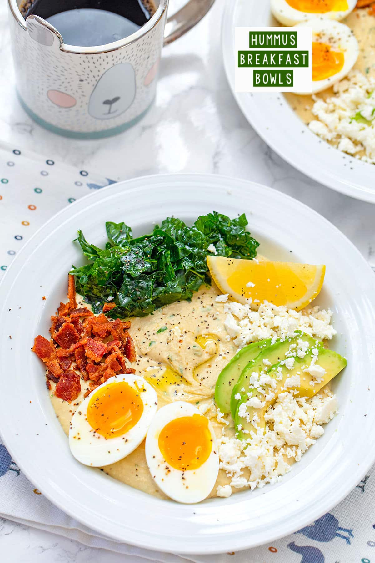 Overhead closeup view of hummus breakfast bowl with halved soft-boiled egg, crumbled bacon, feta cheese, sliced avocado, kale, and lemon wedge with cup of coffee and second hummus bowl in the background with recipe title at top.