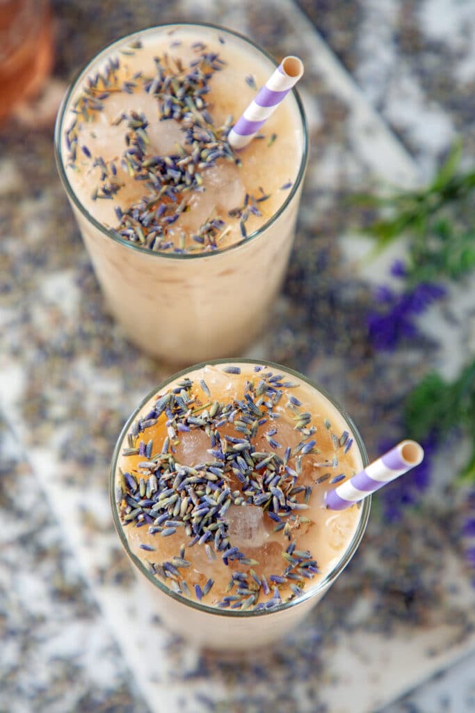 Overhead view of two glasses of iced lavender latte with lavender buds on top and purple and white straws