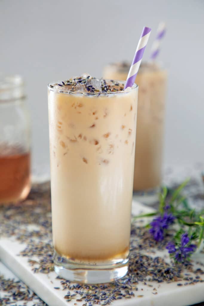 Head-on view of an iced lavender latte in a glass with lavender flowers all around and jar of simple syrup in background