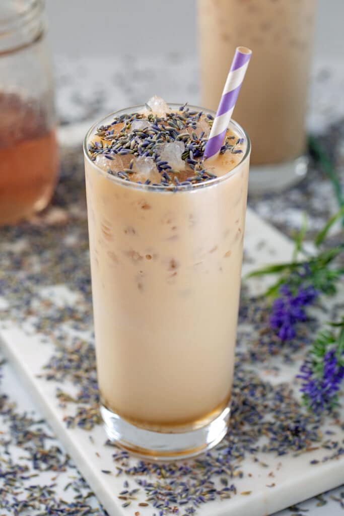 View of a tall glass of an iced lavender latte with lavender buds sprinkled on top and all around and jar of lavender simple syrup in background.