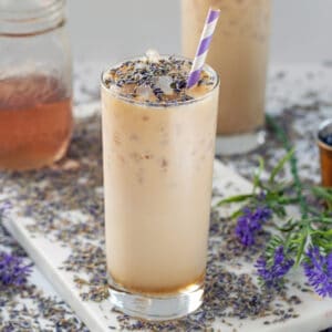 Closeup view of an iced lavender latte topped with dried lavender with more dried lavender all around and jar of lavender simple syrup in background