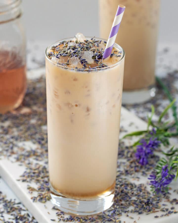 Closeup view of an iced lavender latte topped with dried lavender with more dried lavender all around and jar of lavender simple syrup in background