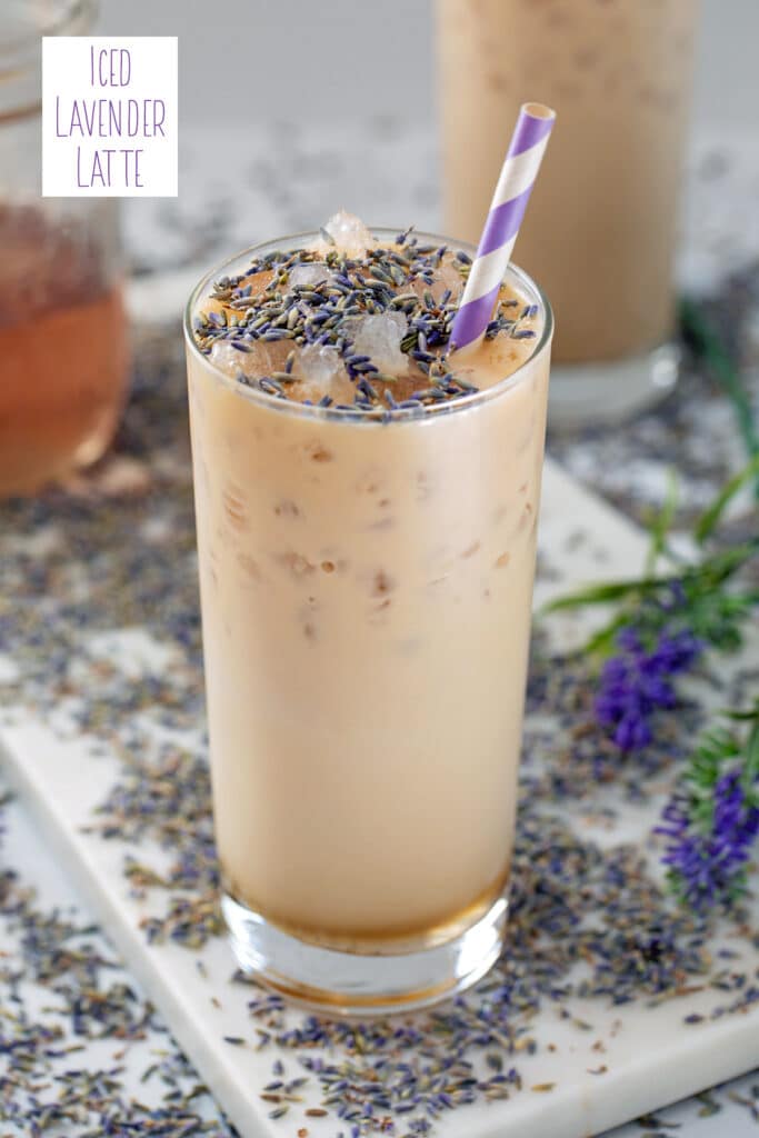 View of a tall glass of an iced lavender latte with lavender buds sprinkled on top and all around and jar of lavender simple syrup in background with recipe title at top