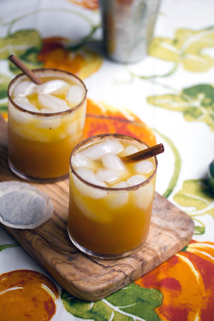 Head-on view of two iced pumpkin green tea cocktails on a wooden board with a tea bag and cocktail shaker in the background