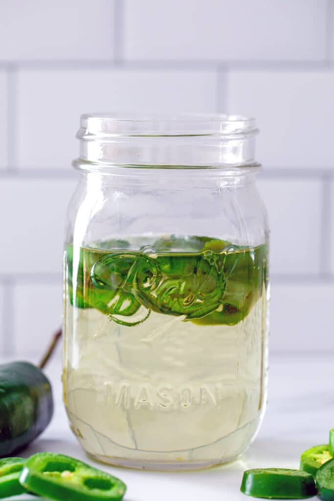 Head-on view of a mason jar filled with jalapeño simple syrup with sliced jalapeños in it and on the table in front